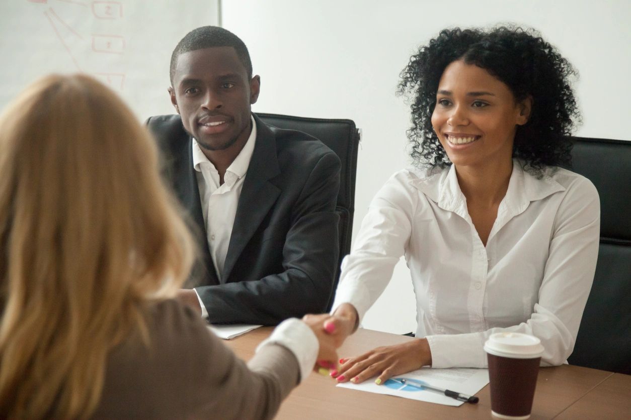 A self-advocate with their back to the viewer sits at a table across from two Black people in a professional setting. The self-advocate and one of the other people are shaking hands to show that people with IDD and employment can work.