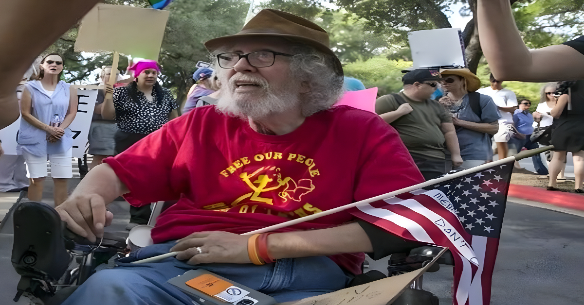 An older man with a hat, glasses, and a gray beard and hair using a wheel chair, holding an American flag, and wearing a t-shirt that say, "Free our people" is in front of a group of people that appear to be at a rally at a capitol building. The image is about remembering Bob Kafka, the man in the photo.
