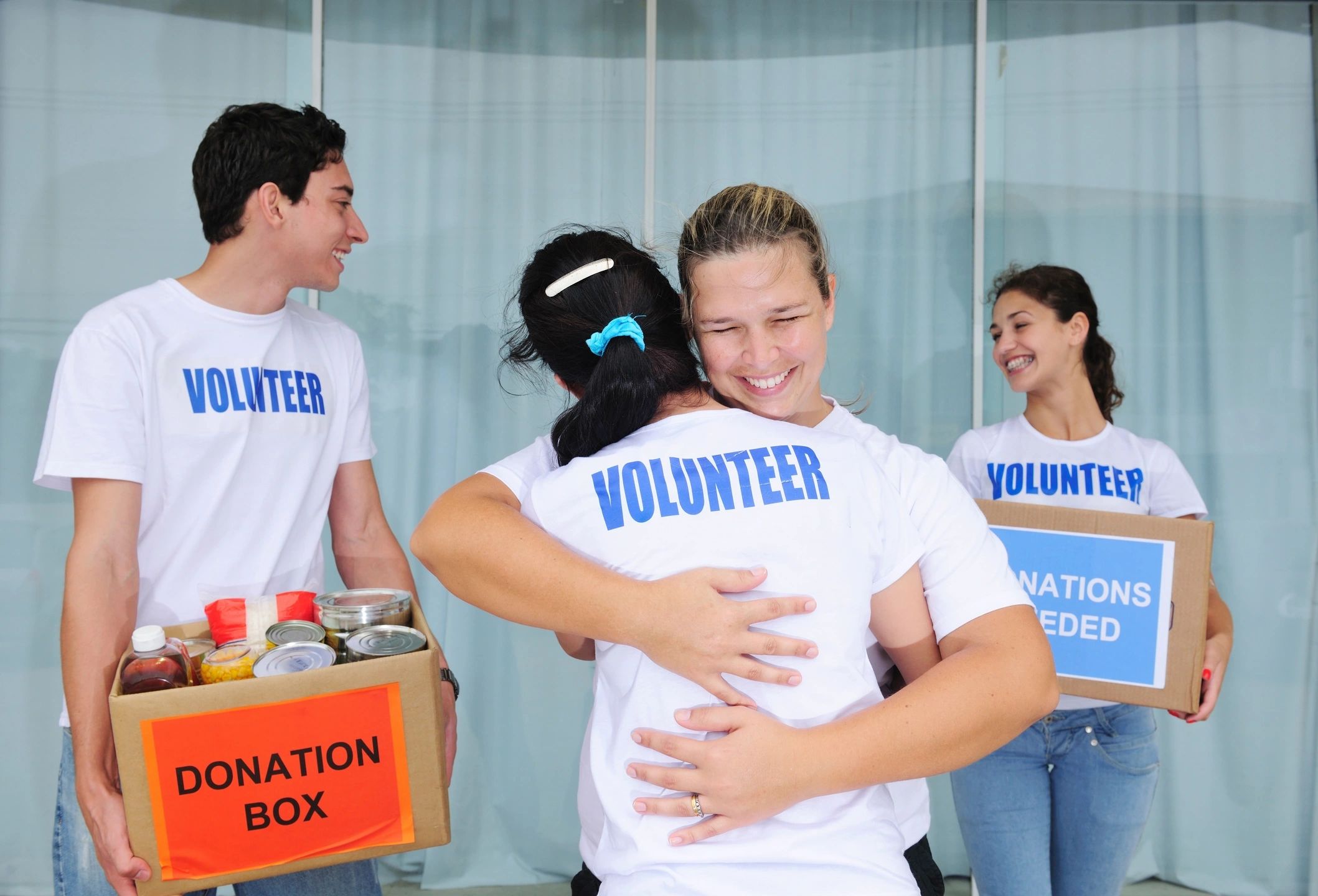 Two people are hugging while two other people are carrying boxes labeled Donation Box. All the people are wearing t-shirts labeled Volunteer. This image reminds the viewer of honoring Dr. King through volunteer service.