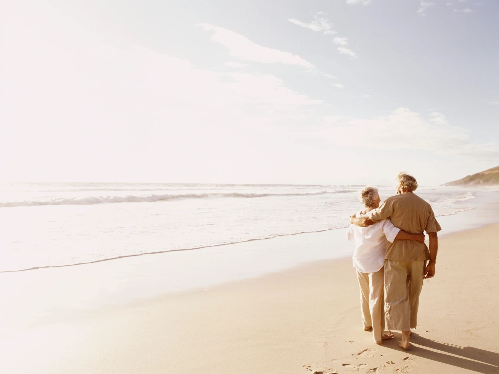 Two people are walking away from the viewer, arm-in-arm on a beach, with the ocean beside them. Seeing the two people together shows the viewer a couple who have shared goals enjoying their time together.