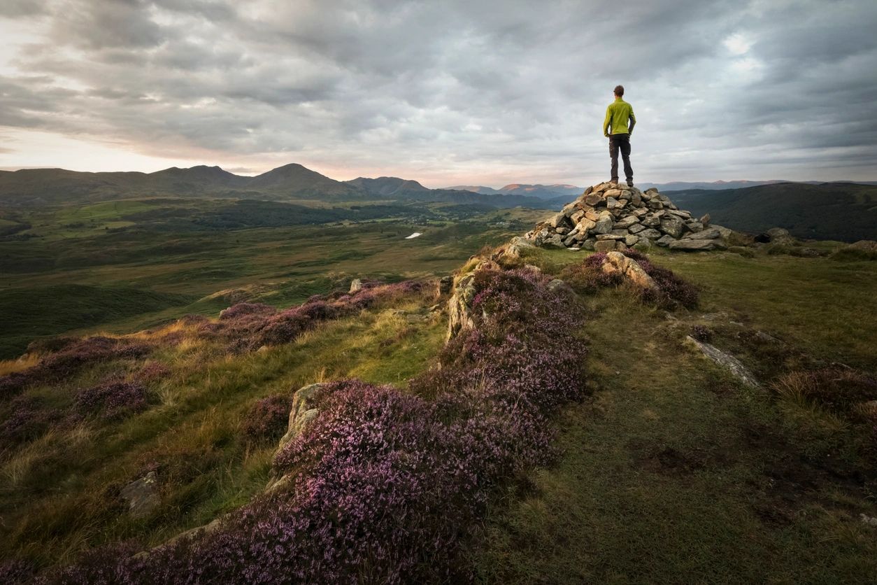 A person stands at the top of a ridge looking over a valley spread out before them with clouds spread across the vast sky. It feels like the person is thinking about what is ahead and coping with change.