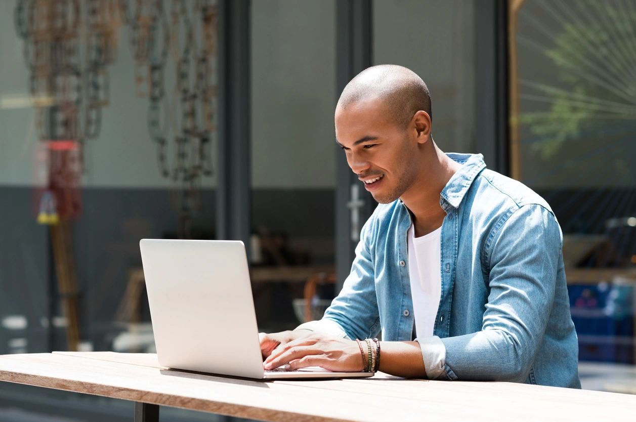 A man sits at a desk working on a laptop computer. The image suggests the man is working on the peer-led program: Real Talk About Independent Living.
