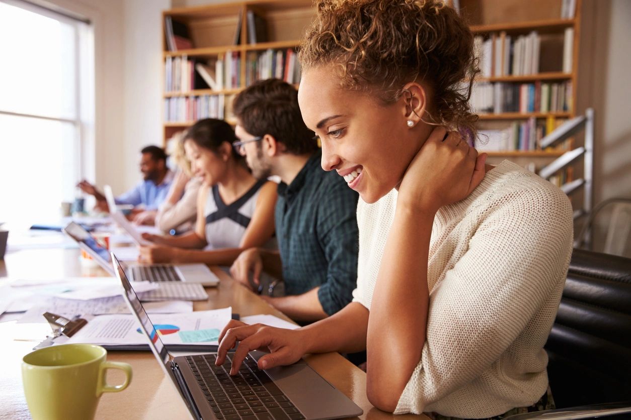 Five people are sitting at a long work table with laptops. They appear to be working together. They are happy building job skills together.